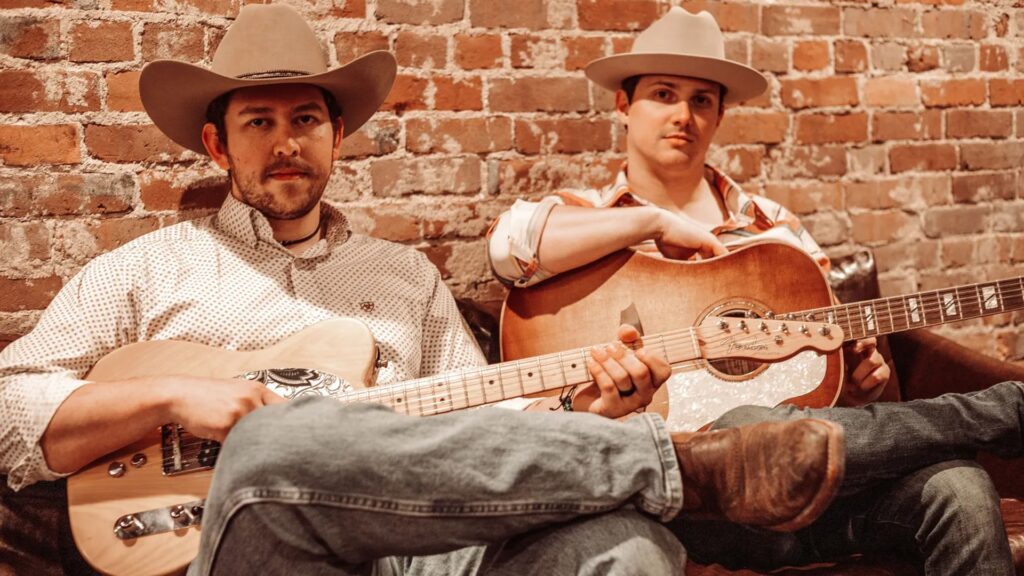 Whisky Outlaws in sepia, seated with guitars against a brick wall, wearing tan cowboy hats.