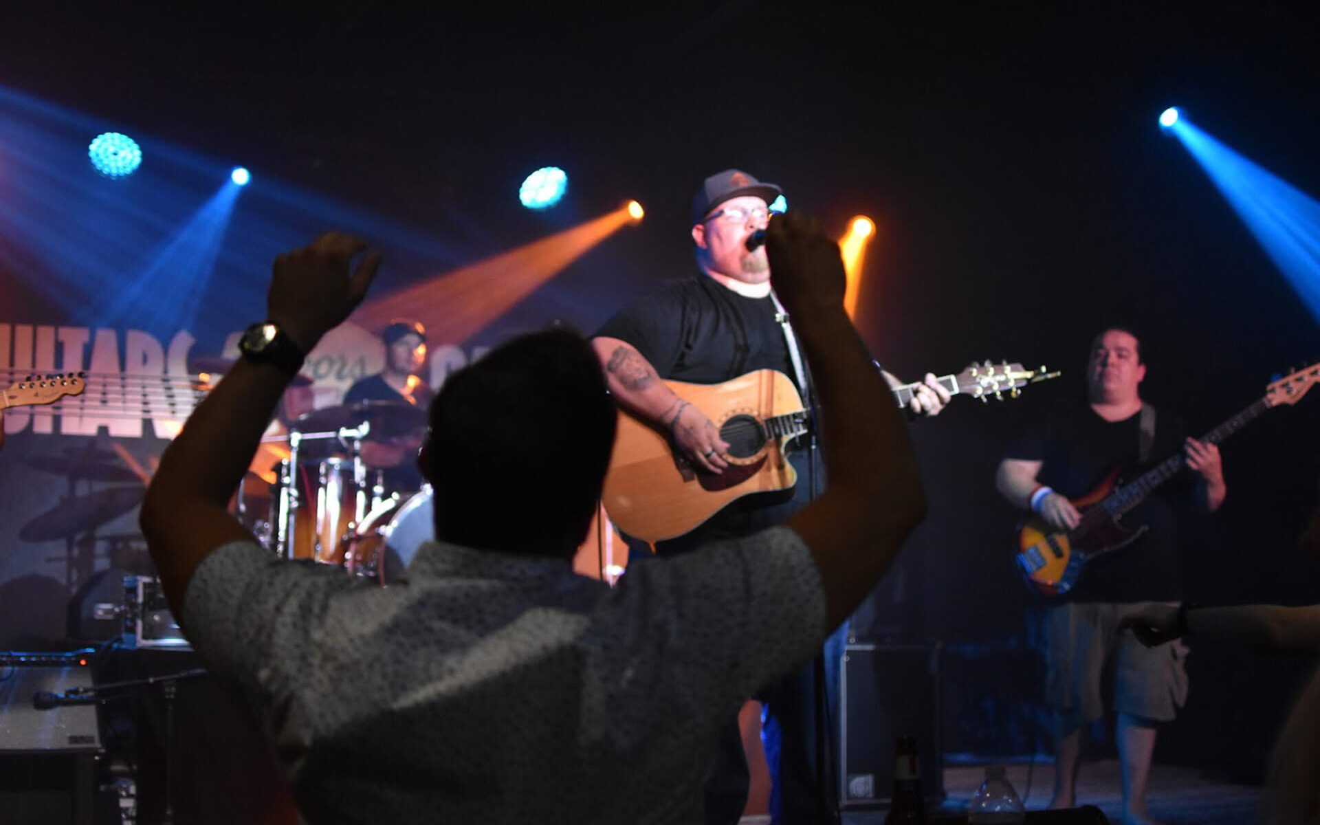 Experience the magic of a Jaron Bell live performance. On a dimly lit stage with vibrant stage lights, Jaron sings into a microphone while playing his guitar. A thrilled fan in the second row raises their hands in joy, surrounded by the band members in the background. The energy of the moment is palpable.